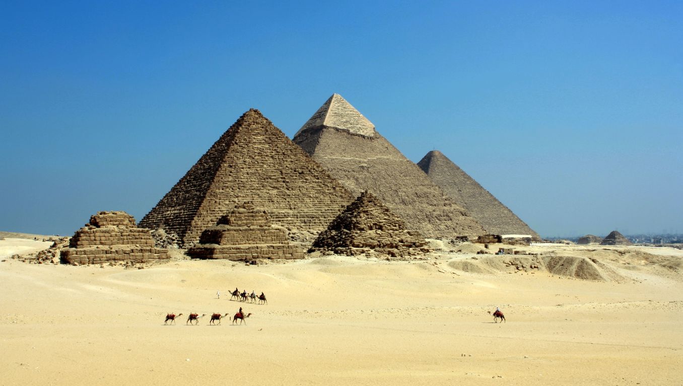 The three Pyramids of Giza seen from the desert plateau with a camel caravan crossing the foreground