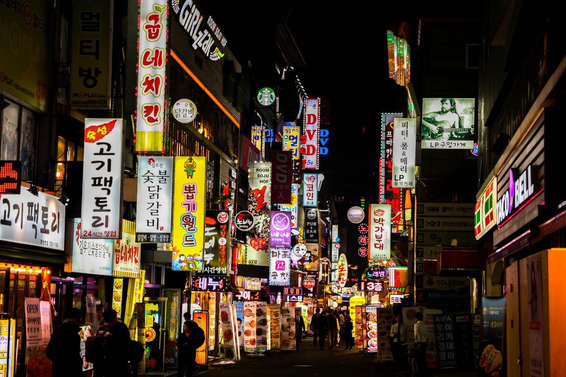 Neon-lit Hongdae street at night with buskers performing under colorful signs and crowds of young Koreans watching